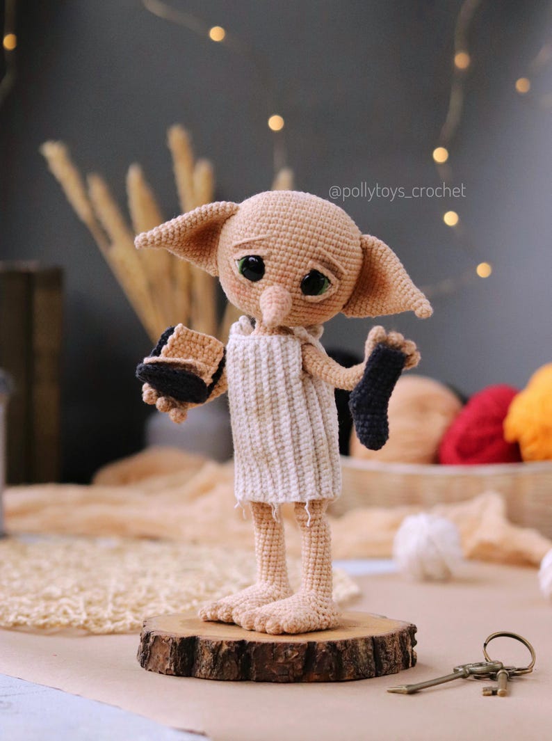 May include: A handmade crochet doll of a character with large ears, green eyes, and a sad expression. The doll wears a white tunic and holds a black sock and a brown object. It stands on a wooden base.