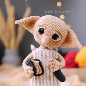 May include: A handmade crochet doll of a character with large ears and green eyes. The doll is dressed in a white and beige striped tunic and holds a book and a dark object. The doll has a sad expression and a whimsical design.