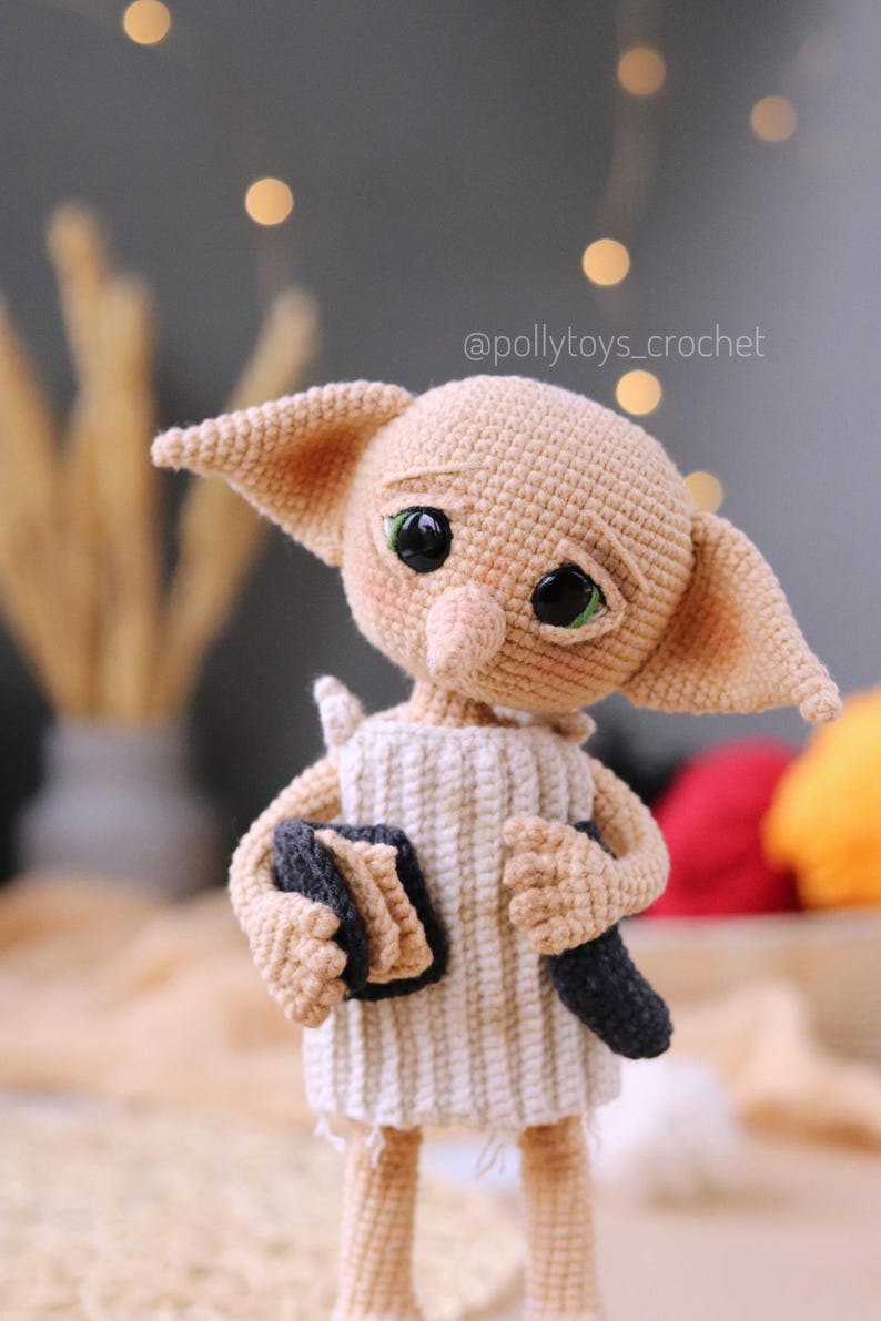 May include: A handmade crochet doll of a character with large ears and green eyes. The doll is dressed in a white and beige striped tunic and holds a book and a dark object. The doll has a sad expression and a whimsical design.