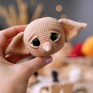 May include: A tan crocheted doll head with large black eyes and green highlights. The doll has large ears and a small nose. The doll's face has a slightly sad expression. The text "@pollytoys_crochet" is visible.
