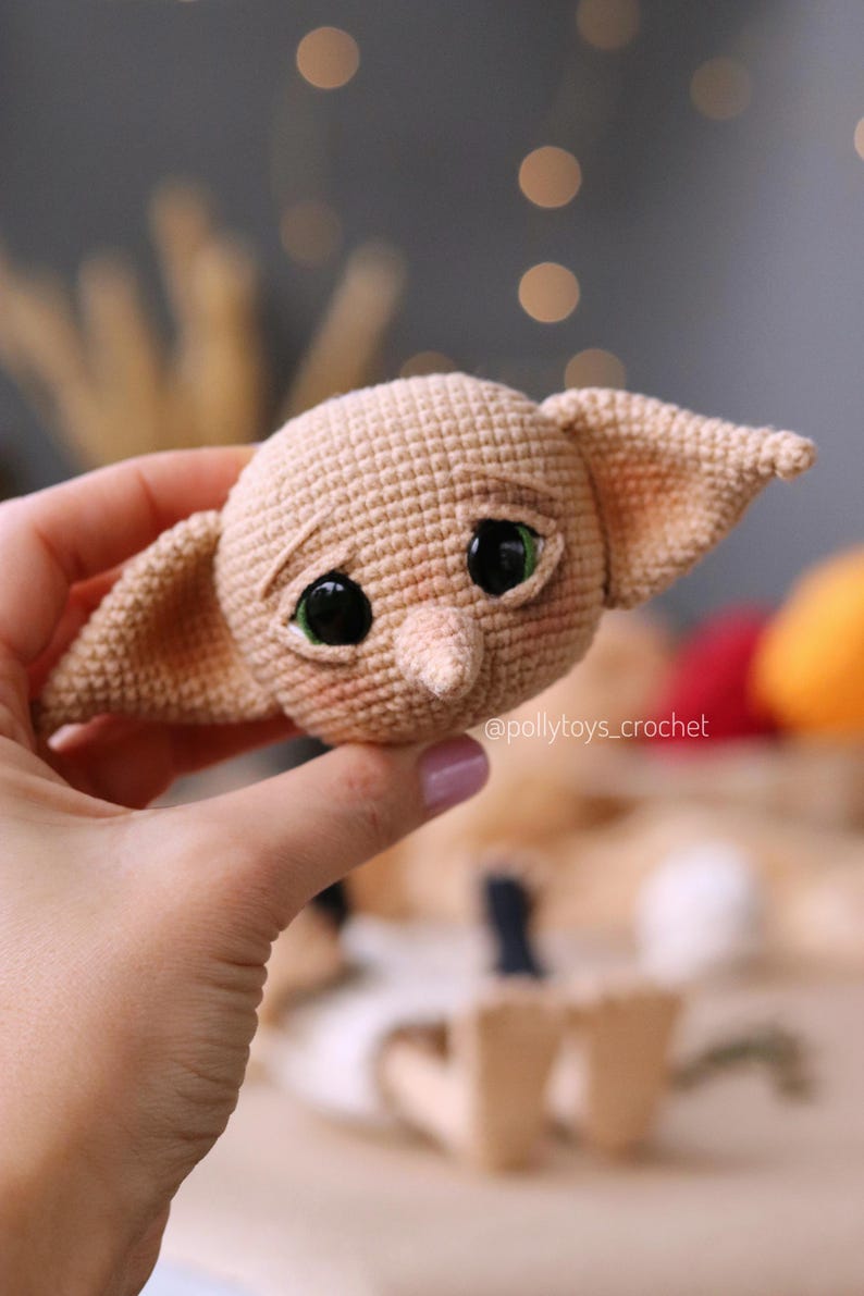 May include: A tan crocheted doll head with large black eyes and green highlights. The doll has large ears and a small nose. The doll's face has a slightly sad expression. The text "@pollytoys_crochet" is visible.