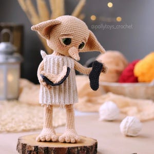 May include: A crocheted doll of a character with large ears, green eyes, and a long nose, wearing a white and beige dress. The doll is holding a black sock. The doll is standing on a wooden slice. The text "@pollytoys_crochet" is visible.