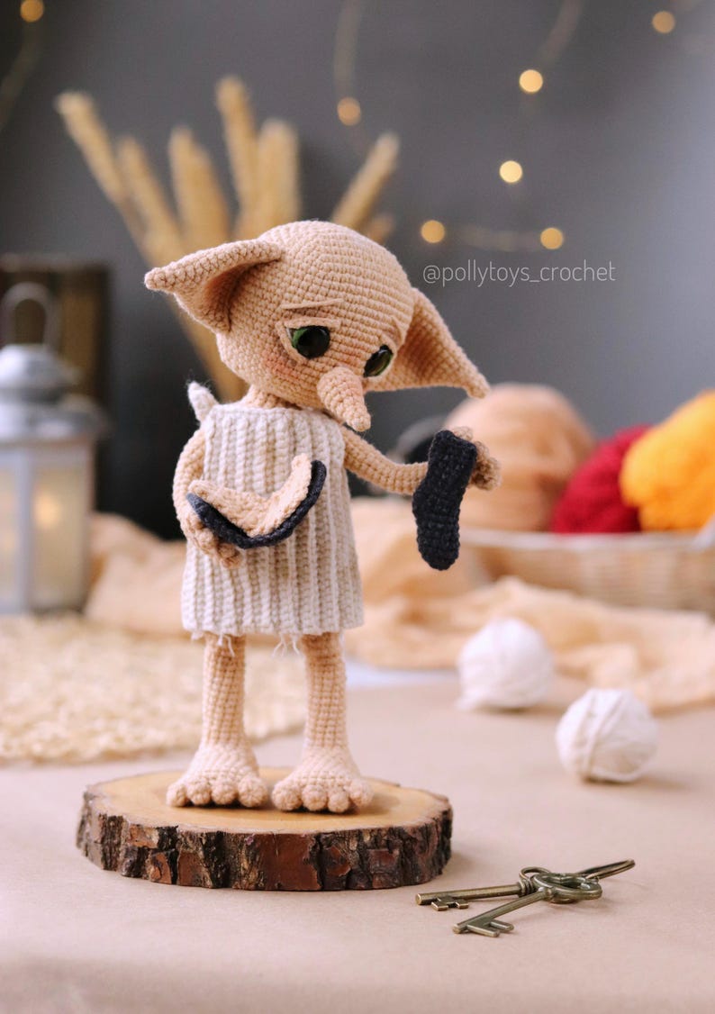 May include: A crocheted doll of a character with large ears, green eyes, and a long nose, wearing a white and beige dress. The doll is holding a black sock. The doll is standing on a wooden slice. The text "@pollytoys_crochet" is visible.