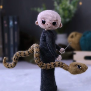 May include: A crocheted amigurumi doll of a bald man wearing a dark grey sweater and holding a silver knife. A brown and gold crocheted snake is wrapped around his legs.