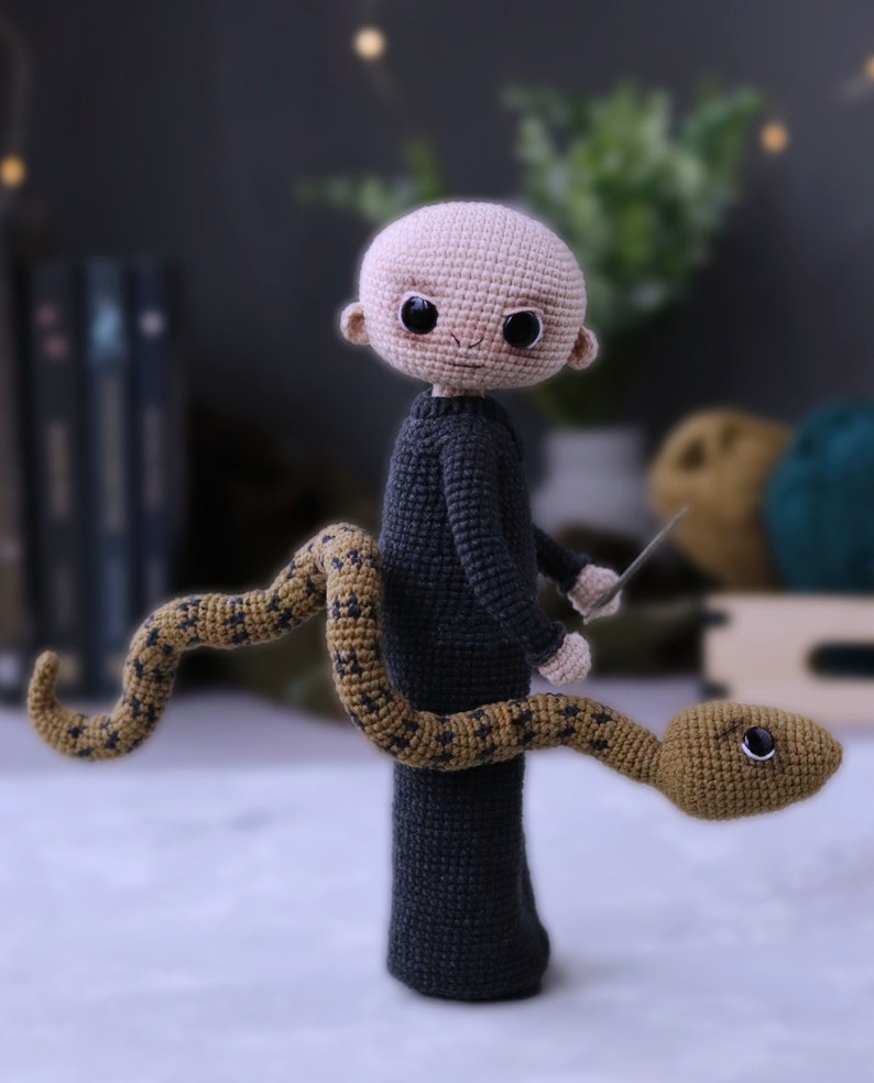 May include: A crocheted amigurumi doll of a bald man wearing a dark grey sweater and holding a silver knife. A brown and gold crocheted snake is wrapped around his legs.