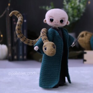 May include: Crocheted amigurumi doll of a young boy with a bald head, wearing a green robe and holding a brown snake. The doll is standing on a white surface. The doll's eyes are black and the snake has a brown and yellow pattern.
