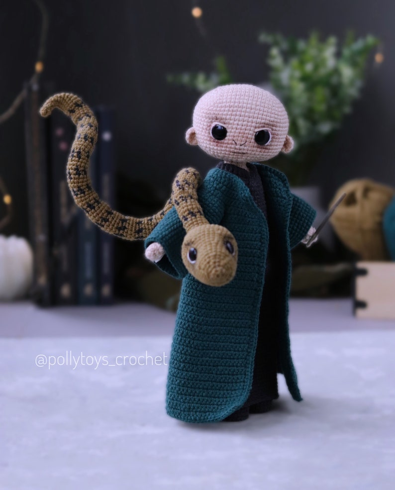 May include: Crocheted amigurumi doll of a young boy with a bald head, wearing a green robe and holding a brown snake. The doll is standing on a white surface. The doll's eyes are black and the snake has a brown and yellow pattern.
