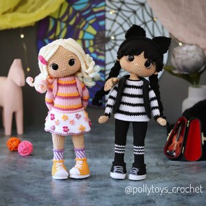 May include: Two crocheted dolls, one with blonde hair wearing a pink and orange striped shirt, a white skirt with pink flowers, and yellow sneakers, and the other with black hair wearing a black and white striped shirt, black pants with white stripes, and black sneakers. The dolls are standing on a gray surface. The text "@pollytoys_crochet" is at the bottom of the image.