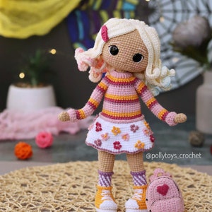 May include: A crocheted doll with blonde hair, wearing a pink and orange striped sweater, a white skirt with pink flowers, and pink and white striped socks. The doll is holding a pink backpack with a heart on it. The doll is standing on a woven mat.