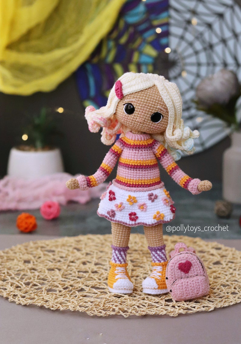 May include: A crocheted doll with blonde hair, wearing a pink and orange striped sweater, a white skirt with pink flowers, and pink and white striped socks. The doll is holding a pink backpack with a heart on it. The doll is standing on a woven mat.