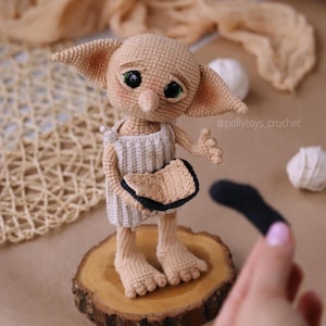 May include: A crocheted doll of a fictional character with large ears, green eyes, and a neutral skin tone. The doll wears a white and beige outfit and stands on a wooden slice. A black sock is held nearby. The doll is a whimsical, handcrafted item.