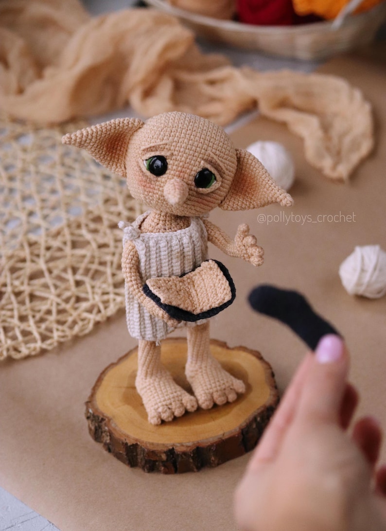 May include: A crocheted doll of a fictional character with large ears, green eyes, and a neutral skin tone. The doll wears a white and beige outfit and stands on a wooden slice. A black sock is held nearby. The doll is a whimsical, handcrafted item.