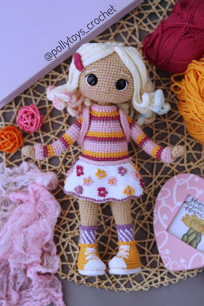 May include: A crocheted doll with blonde hair, wearing a pink and yellow striped shirt, a white skirt with pink flowers, and yellow and white sneakers. The doll is sitting on a woven mat.