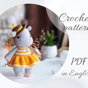 Puede incluir: Patrón de crochet PDF en inglés para un hipopótamo de crochet gris que lleva un sombrero marrón, un vestido amarillo con rayas blancas y una cinta amarilla.