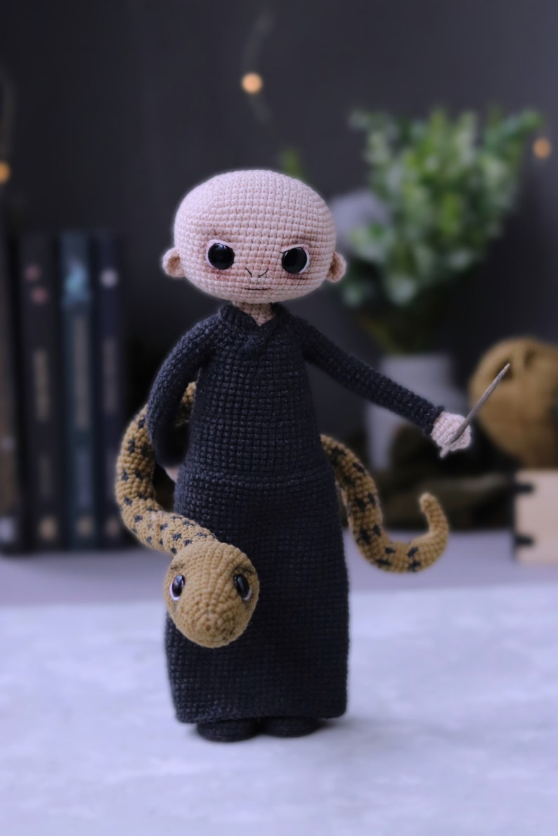 May include: A crocheted doll with a bald head, wearing a black robe and holding a snake and a wand. The doll has a friendly expression with large eyes and a small smile.