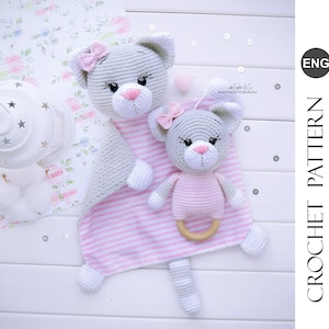 Puede incluir: Un juguete de gato de crochet gris con un lazo rosa y un juguete de gato de crochet gris más pequeño con un lazo rosa y una manta a rayas rosa y blanca. El juguete de gato más grande tiene una cola y el juguete de gato más pequeño tiene un anillo de madera unido.