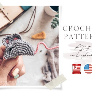 May include: Crochet pattern for a grey mouse with pink ears. The pattern is available in a PDF format in English. The image shows a crocheted mouse being held in a hand.