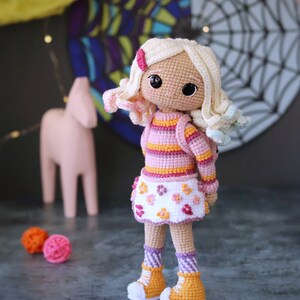 May include: A crocheted doll with blonde hair, wearing a pink and orange striped sweater, a white skirt with pink flowers, and yellow and white shoes. The doll is standing on a gray surface.
