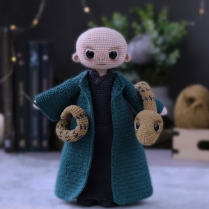 May include: A crocheted doll with a bald head, wearing a dark green robe and holding a crocheted snake. The doll has large eyes and a small mouth.