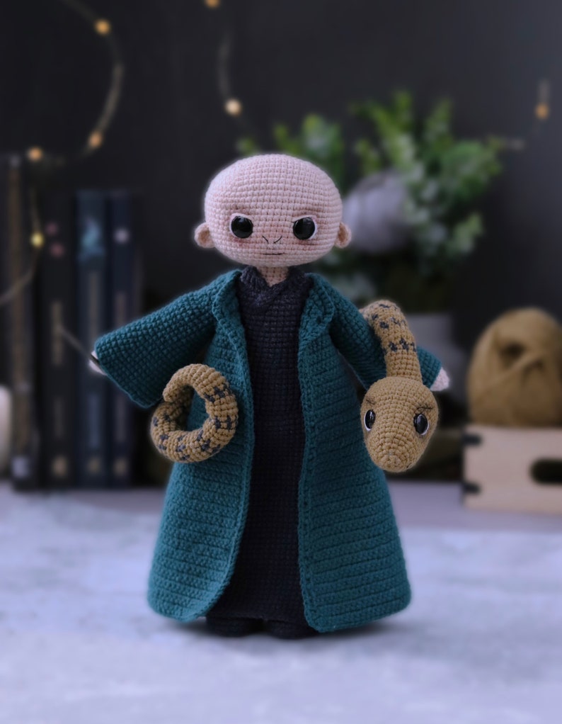 May include: A crocheted doll with a bald head, wearing a dark green robe and holding a crocheted snake. The doll has large eyes and a small mouth.