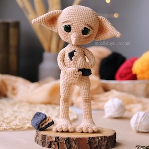 May include: A handmade crocheted doll of a character with large ears and big eyes. The doll is beige and stands on a small wooden base. A small hat is next to the doll. The doll has a black patch on its chest.