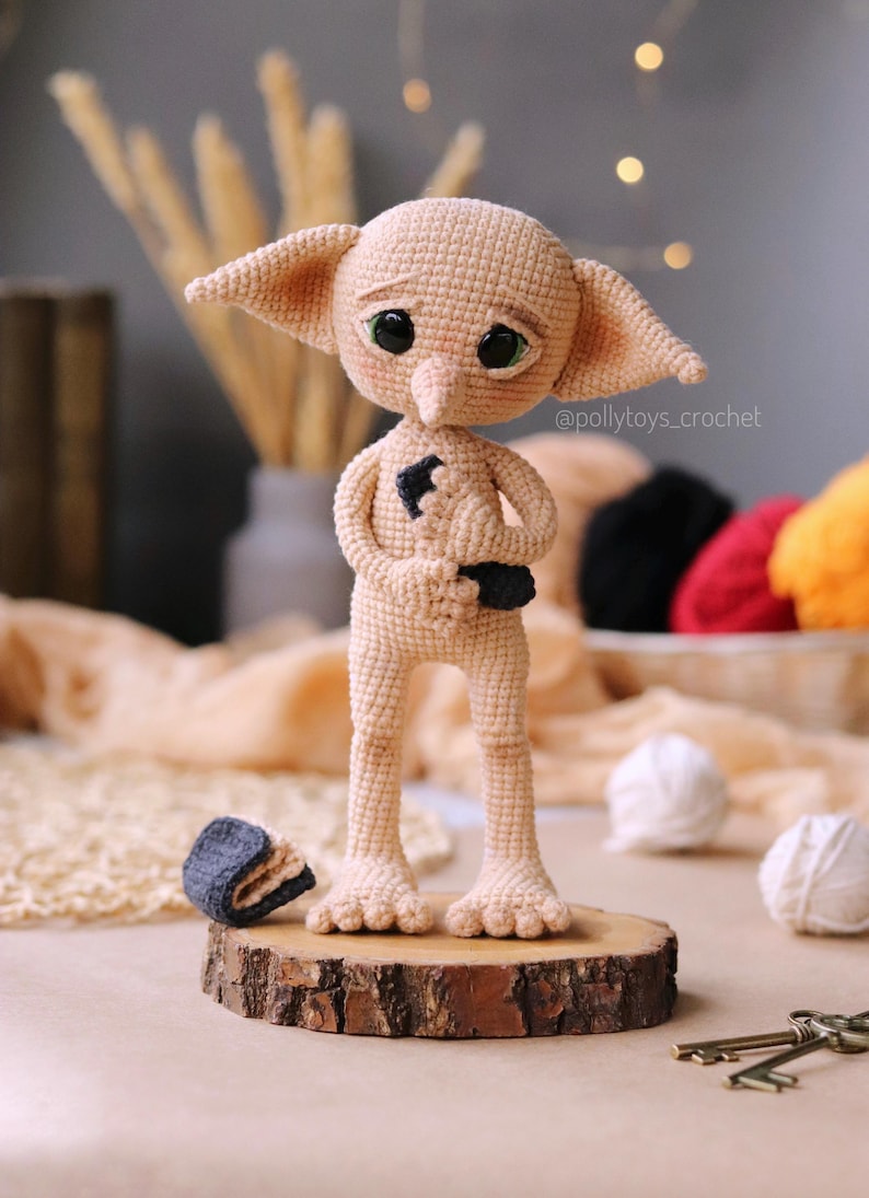 May include: A handmade crocheted doll of a character with large ears and big eyes. The doll is beige and stands on a small wooden base. A small hat is next to the doll. The doll has a black patch on its chest.