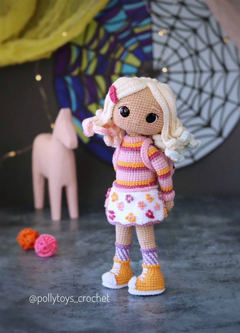 May include: A crocheted doll with blonde hair, wearing a pink and orange striped sweater, a white skirt with pink flowers, and yellow and white shoes. The doll is standing on a gray surface.