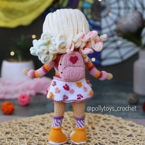 May include: A crocheted doll wearing a pink and white dress, a pink backpack with a heart on it, and orange and white shoes. The doll has blonde hair with pink and blue accents. The doll is standing on a woven mat. @pollytoys_crochet