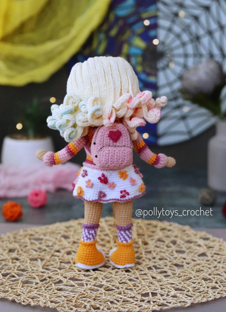 May include: A crocheted doll wearing a pink and white dress, a pink backpack with a heart on it, and orange and white shoes. The doll has blonde hair with pink and blue accents. The doll is standing on a woven mat. @pollytoys_crochet