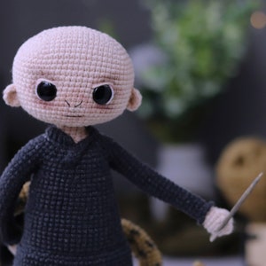 May include: A crocheted doll with a bald head, black eyes, and a black sweater. The doll is holding a wooden stick in its right hand.