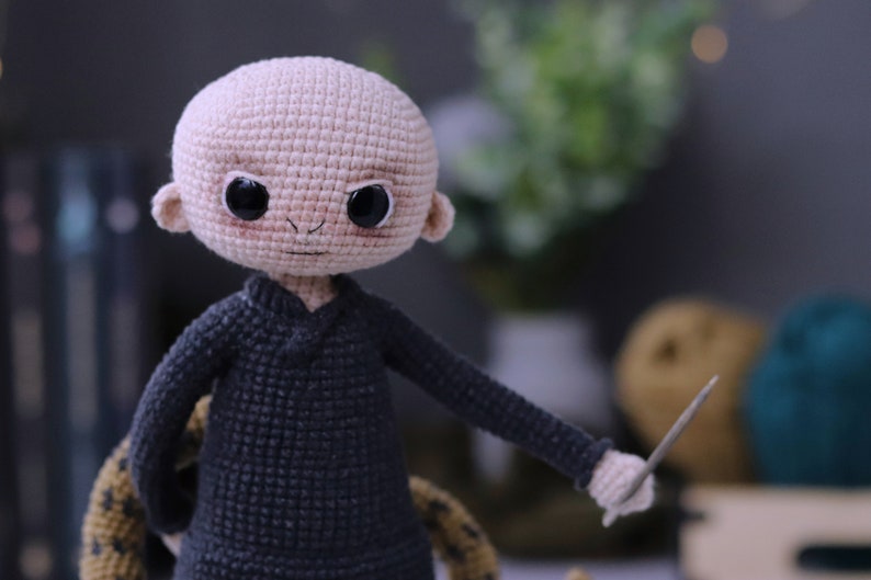 May include: A crocheted doll with a bald head, black eyes, and a black sweater. The doll is holding a wooden stick in its right hand.