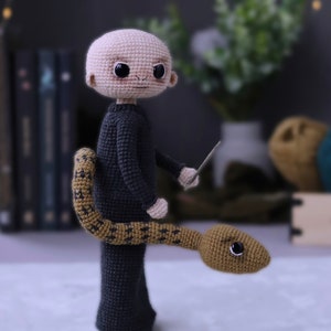 May include: A crocheted plush toy of a bald man wearing a black robe and holding a wand. A brown snake with black markings is wrapped around his legs.