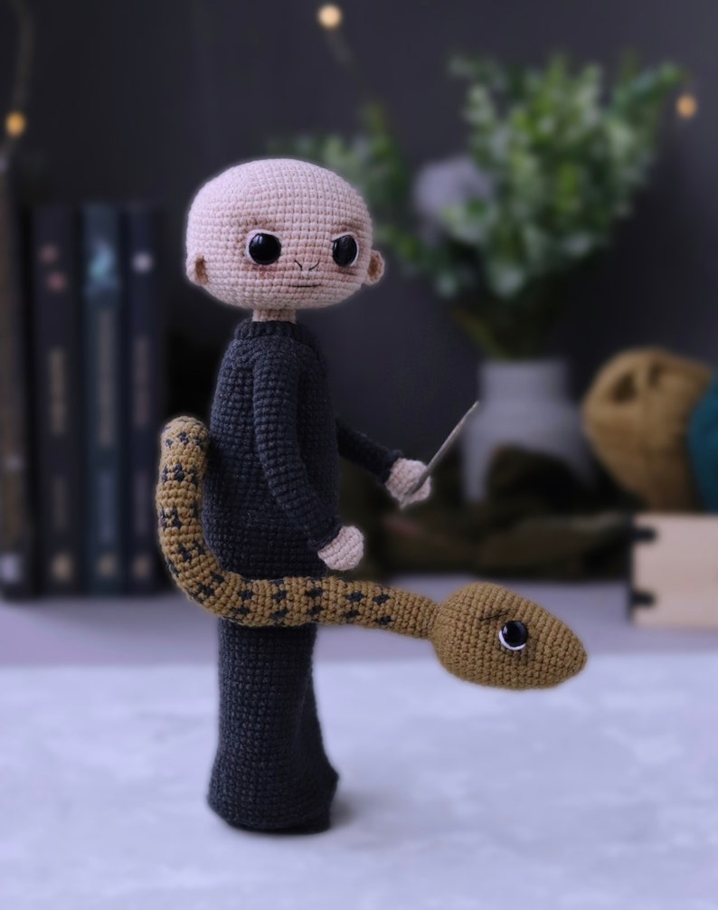 May include: A crocheted plush toy of a bald man wearing a black robe and holding a wand. A brown snake with black markings is wrapped around his legs.