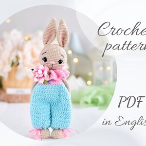May include: Crochet pattern PDF in English for a beige bunny wearing blue overalls and holding a pink flower.