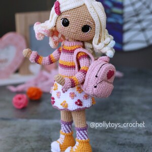 May include: A crocheted doll with blonde hair, wearing a pink and orange striped sweater, a white skirt with pink flowers, pink and white striped socks, and yellow shoes. The doll is carrying a pink backpack with a heart on it. The doll is standing on a gray surface. @pollytoys_crochet