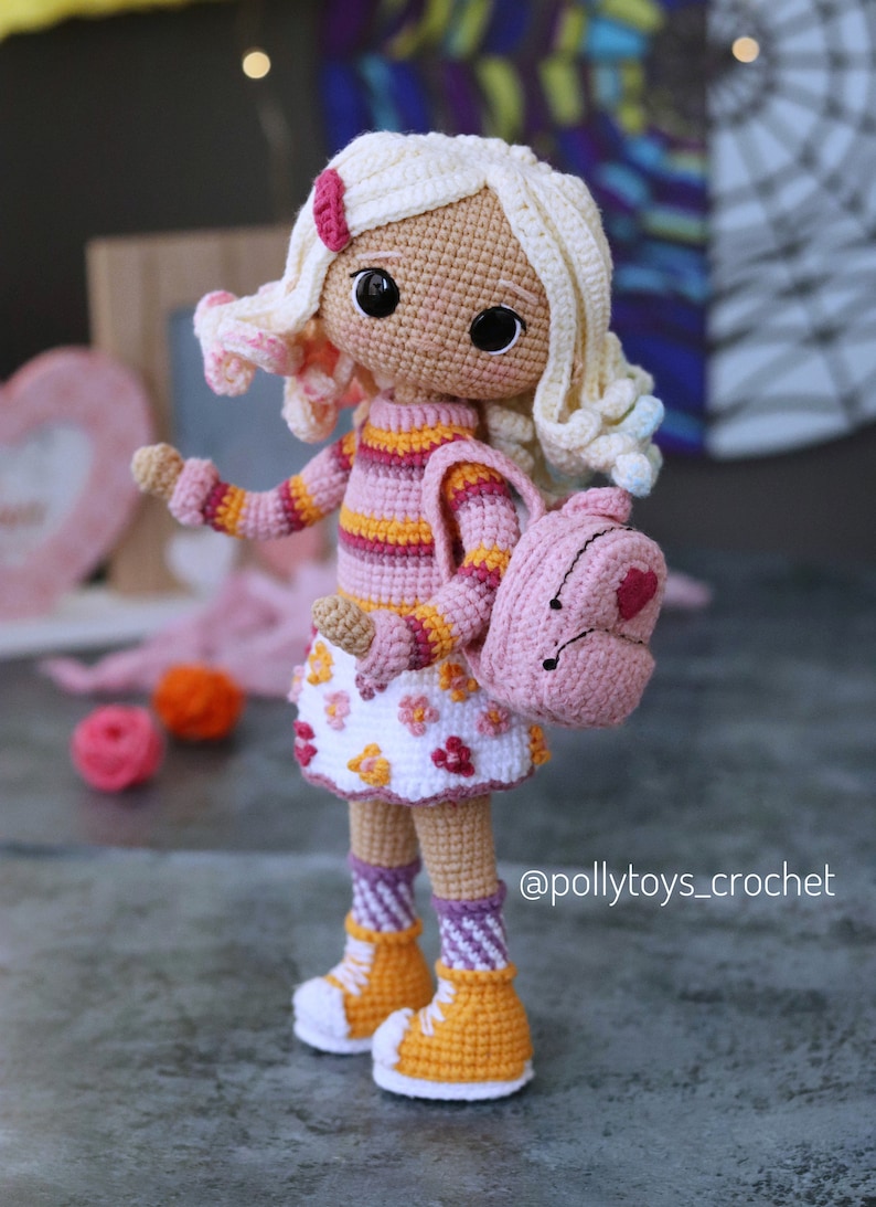 May include: A crocheted doll with blonde hair, wearing a pink and orange striped sweater, a white skirt with pink flowers, pink and white striped socks, and yellow shoes. The doll is carrying a pink backpack with a heart on it. The doll is standing on a gray surface. @pollytoys_crochet
