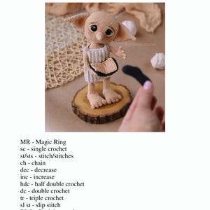 May include: A handmade crochet doll of a character with large ears, wearing a beige outfit, and holding a black object. The doll is displayed on a wooden slice. The image includes the text "Abbreviations".