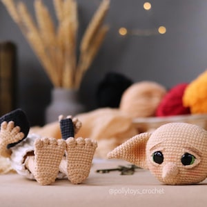 May include: A handmade crochet doll of a character with large ears and big eyes, lying on a table. The doll is beige with black accents. A ball of yarn and other crochet items are in the background.