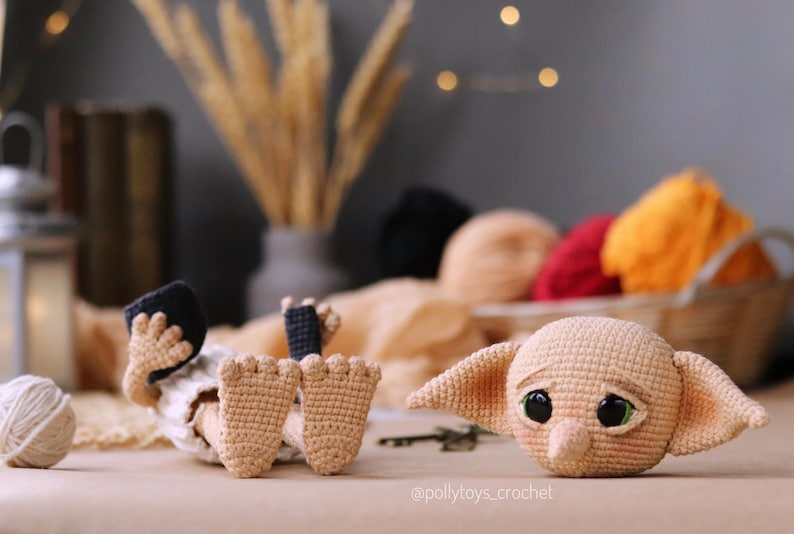 May include: A handmade crochet doll of a character with large ears and big eyes, lying on a table. The doll is beige with black accents. A ball of yarn and other crochet items are in the background.