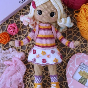 May include: A crocheted doll with blonde hair, wearing a pink and yellow striped shirt, a white skirt with pink flowers, and yellow and white sneakers. The doll is sitting on a woven mat.