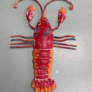May include: A red and orange metal lobster wall decoration. The lobster has curved antennae, claws, and multiple legs. The body is segmented with orange and red paint. The lobster is a decorative item for a coastal or nautical theme.