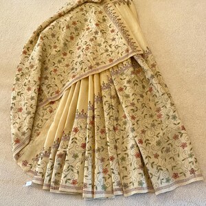 May include: Cream-colored textile with intricate floral embroidery. The design features delicate flowers in shades of red, green, and pink, along with swirling vines. The edges are finished with a detailed border. The fabric is draped, showcasing the detailed pattern.