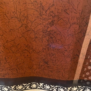 May include: A sheer fabric with a brown background and dark brown, abstract, linear patterns. The bottom edge features a black border with a cream-colored floral design. A vertical stripe of light brown is visible on the right side.