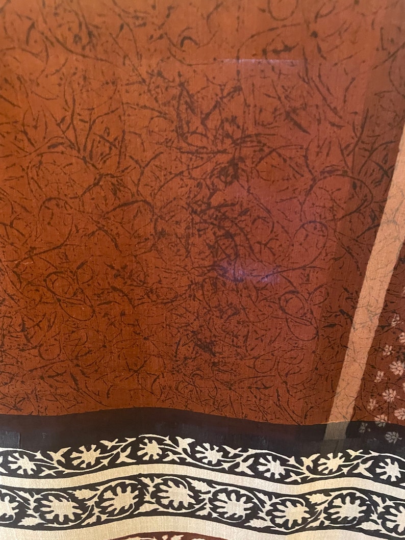 May include: A sheer fabric with a brown background and dark brown, abstract, linear patterns. The bottom edge features a black border with a cream-colored floral design. A vertical stripe of light brown is visible on the right side.