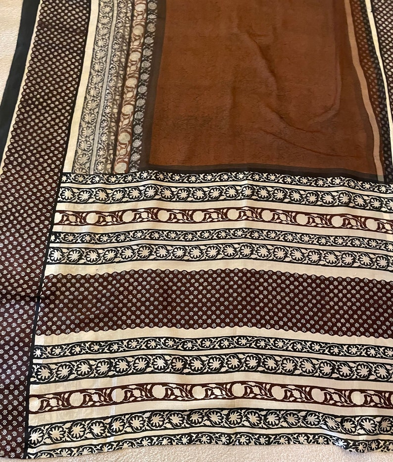 May include: A brown and cream-colored textile with intricate patterns. The design includes floral and geometric motifs in black and brown, with a dark brown central panel. The edges feature a dotted pattern.