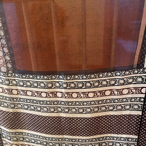 May include: A sheer brown curtain with a dark brown border. The curtain has a patterned design in white and dark brown. The pattern includes floral and geometric elements. The curtain is hanging in front of a window.
