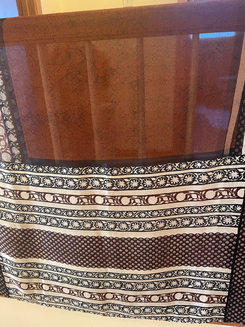 May include: A sheer brown curtain with a dark brown border. The curtain has a patterned design in white and dark brown. The pattern includes floral and geometric elements. The curtain is hanging in front of a window.