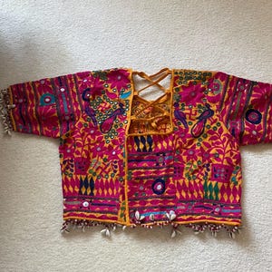 May include: A vibrant, embroidered jacket in shades of pink, yellow, and blue. The jacket features intricate patterns, mirror accents, and shell embellishments along the sleeves and hem. The back has a crisscross tie closure.