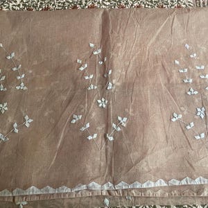 May include: A sheer, light brown fabric textile featuring three repeating embroidered floral motifs in white thread. The fabric has a scalloped white border along the bottom edge, with delicate leaf and petal patterns stitched across the surface.