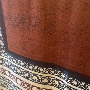 May include: A brown textile with a dark pattern and a decorative border. The border features a repeating floral design in cream and brown against a black background. The main section has a textured appearance.